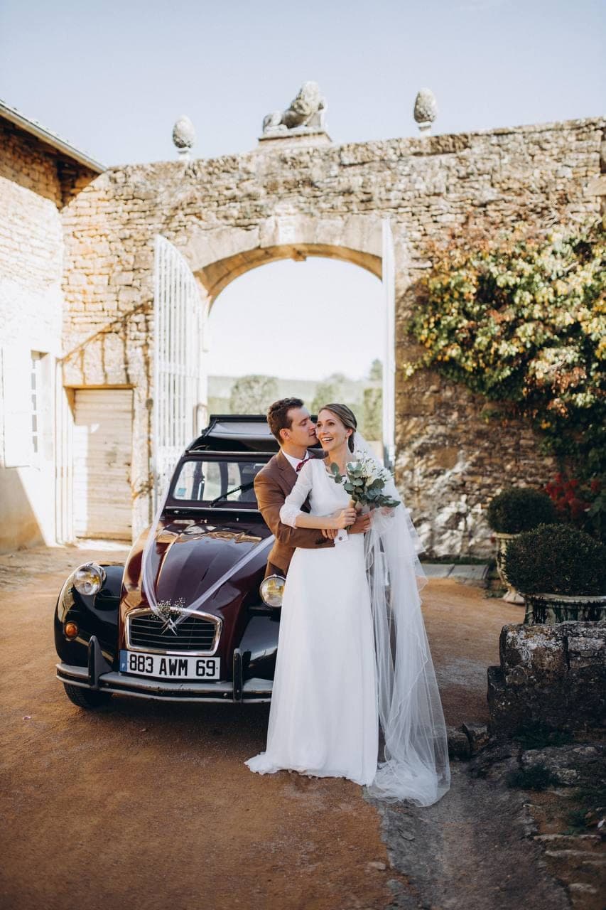 Les mariés devant une 2CV bordeaux, à l'entrée d'un château.