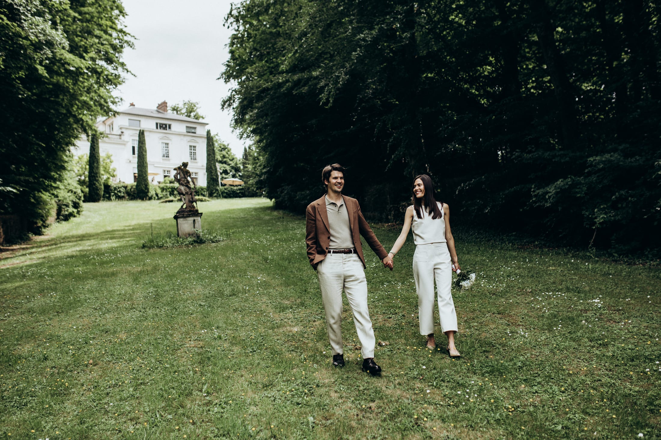 A couple walks side by side across a wide lawn, a mansion in the distance.